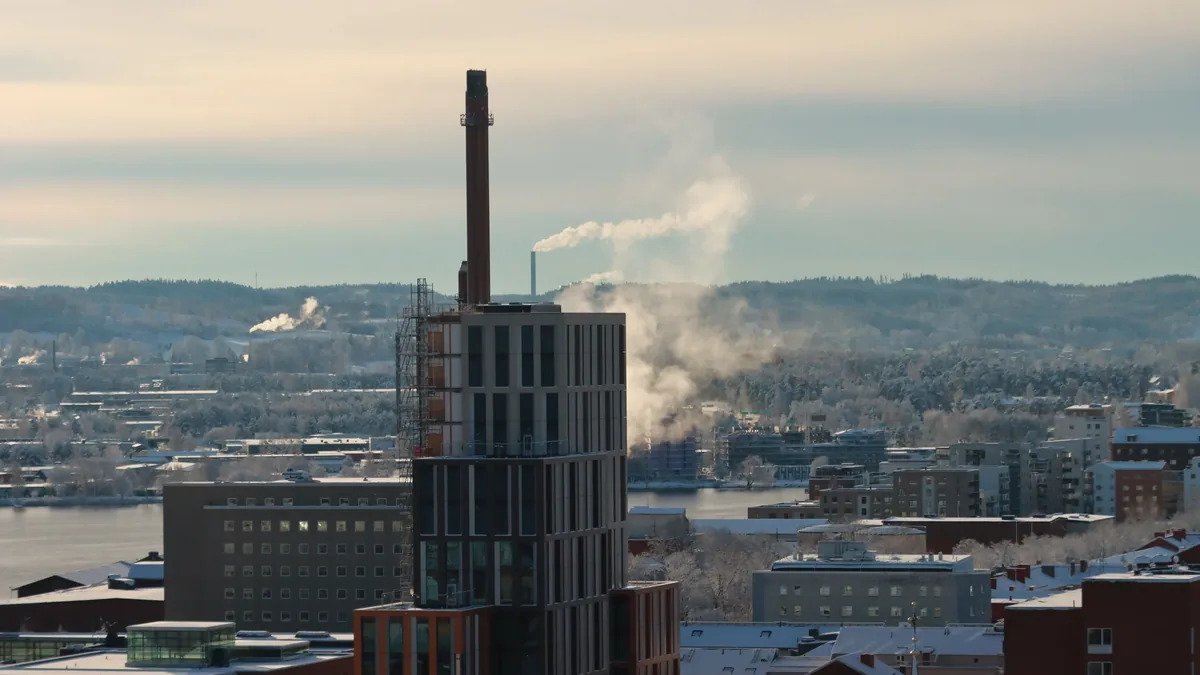 A cityscape with a prominent smokestack emitting smoke, set against a backdrop of buildings and a body of water.