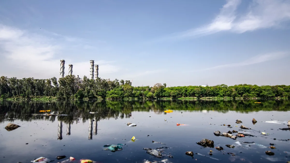 A polluted body of water with scattered trash and debris, set against a backdrop of trees and a blue sky.