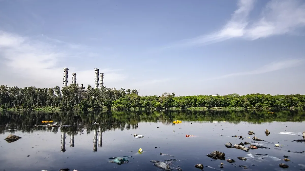 A polluted body of water with scattered trash and debris, set against a backdrop of trees and industrial structures.
